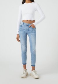 Jean skinny bleu clair avec des détails déchirés et une taille haute. Associé à un haut blanc à manches longues court et des baskets blanches.