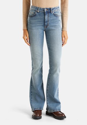 Lichtblauwe high-waist flared jeans gecombineerd met bruine schoenen met een stevige zool en een beige top met lange mouwen, getoond van de taille tot de voeten.