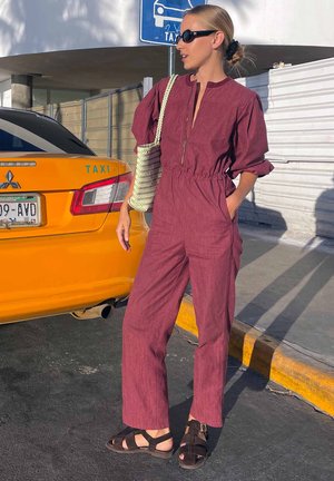 Woman wearing a maroon jumpsuit and black sandals stands on street near yellow taxi, carrying a woven shoulder bag and wearing black sunglasses.