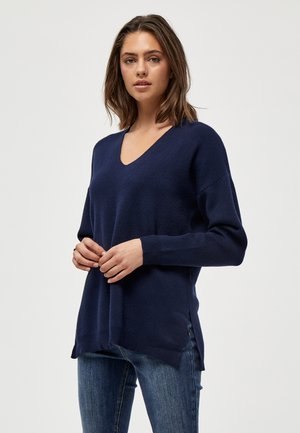 Navy blue knit sweater with a V-neck, long sleeves, and side slits. Soft texture, casual fit. Worn with dark blue jeans.