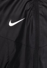 Černá větrovka Nike s bílým logem, hladkou texturou, minimalistickým designem a středovým zipem. Materiál je lehký a odolný.