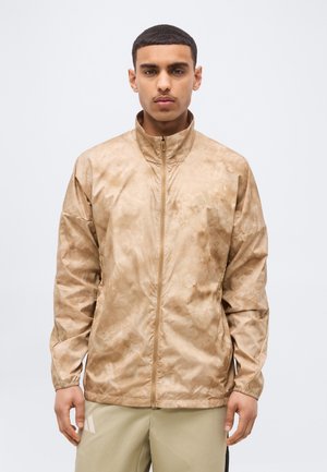 Ung mann iført en beige mønstret jakke med glidelås og lyse bukser, stående mot en ensfarget lys grå bakgrunn.