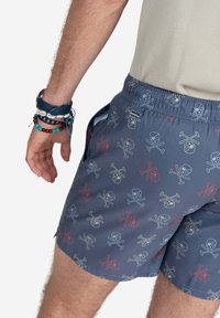 Bañador azul con un divertido patrón de calaveras y tibias en varios colores. Cintura elástica y bolsillos laterales. Tela suave y ligera.