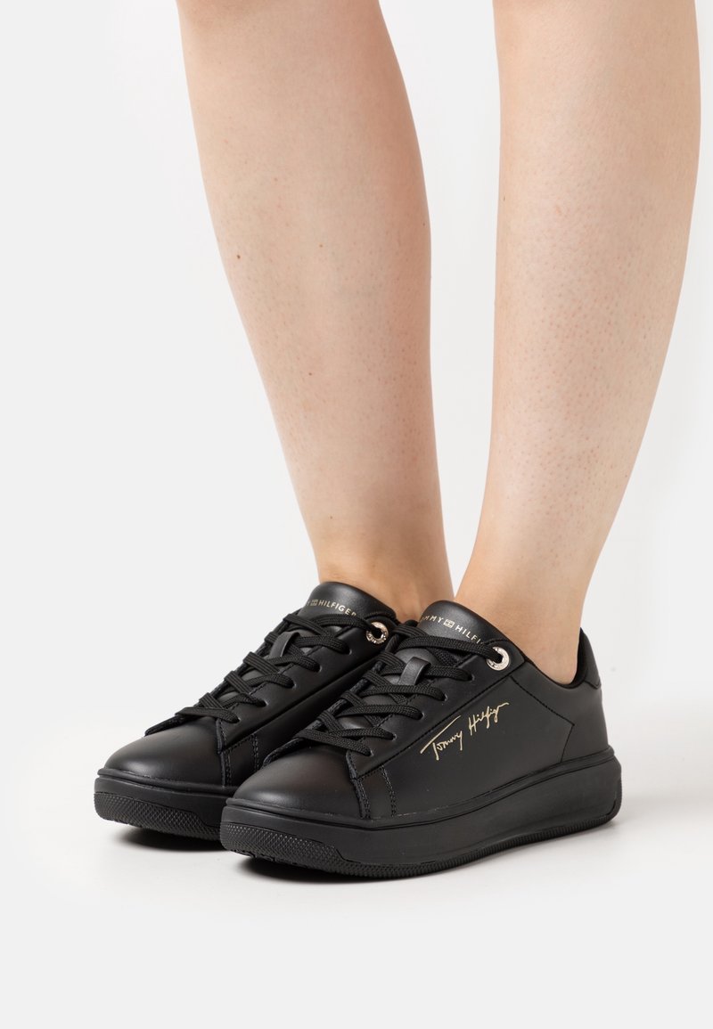 Baskets basses noires avec lacets noirs portées aux pieds, avec la signature dorée "Tommy Hilfiger" sur le côté et le nom de la marque sur la languette.