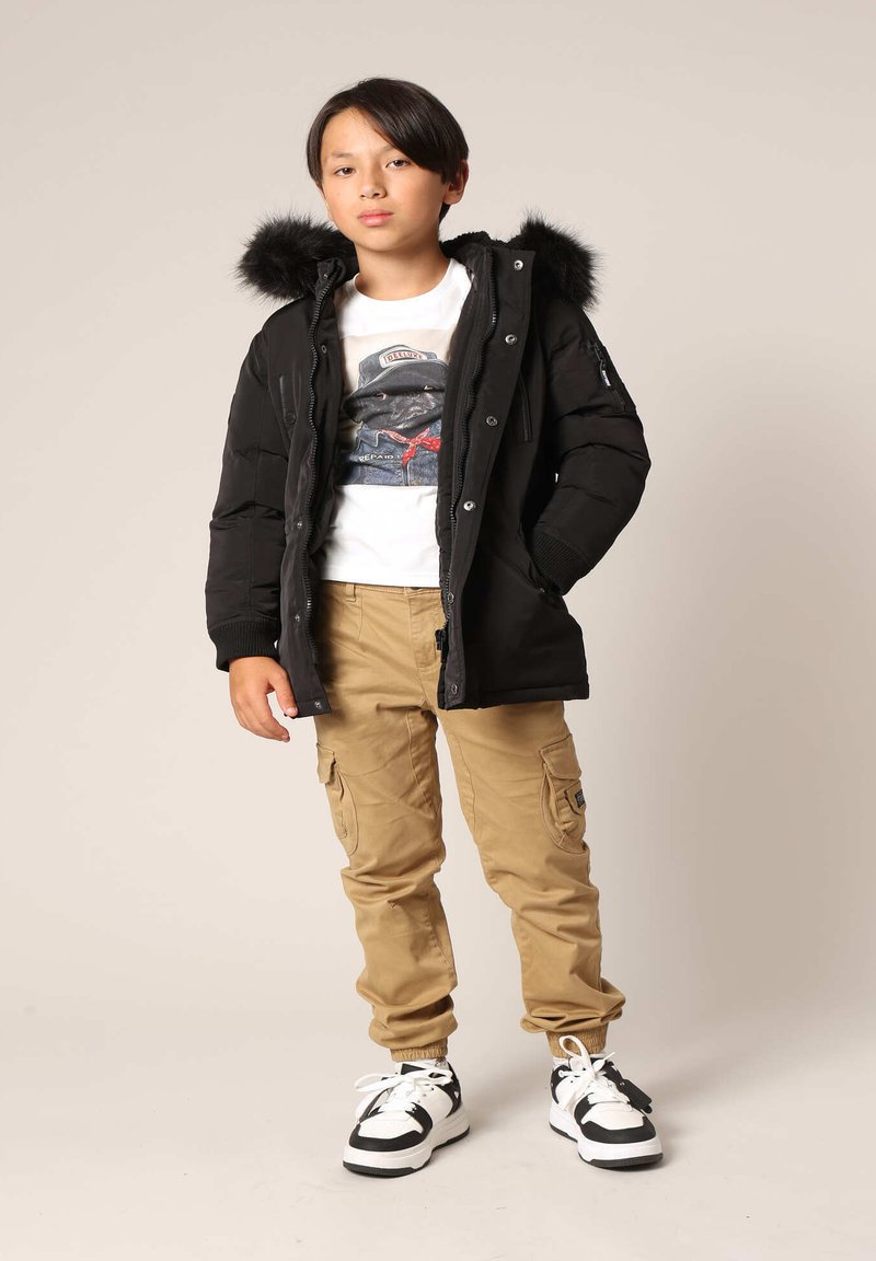 Veste matelassée noire avec capuche en fourrure, associée à un t-shirt graphique blanc, un pantalon cargo kaki et des baskets noires et blanches.