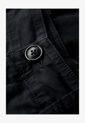 Tessuto nero con un grande bottone nero, caratterizzato da un design a quattro fori. Texture liscia con dettagli di cucitura visibili nei dintorni.