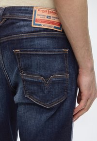 Σκούρα μπλε τζιν από denim με ραφές στην πίσω τσέπη και δερμάτινη ετικέτα Diesel Industry στην μέση, φορεμένα από ένα άτομο με ορατό χέρι.