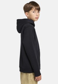 Sudadera de algodón negra con un bolsillo frontal y puños acanalados, vista desde un lado, combinada con pantalones de color claro. Textura suave.