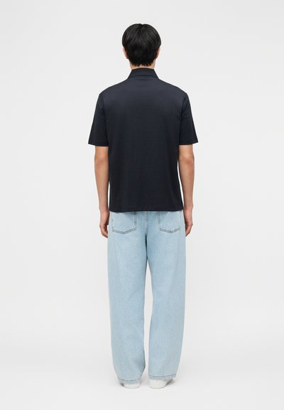 Άνδρας που στέκεται με την πλάτη στην κάμερα, φορώντας μαύρο μπλουζάκι με κοντά μανίκια, ανοιχτόχρωμο μπλε τζιν με φαρδιά παντελόνια και λευκά παπούτσια σε απλό φόντο.