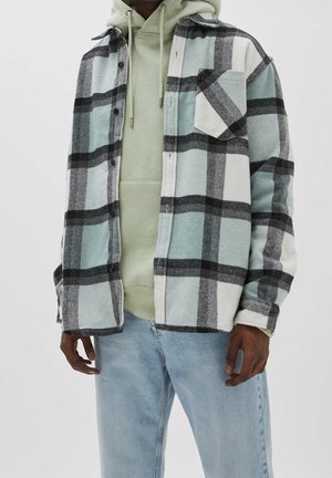 Personne portant un sweat à capuche vert clair sous une chemise surdimensionnée à carreaux ouverte aux motifs noirs, blancs et bleu pastel, associée à un jean bleu clair.