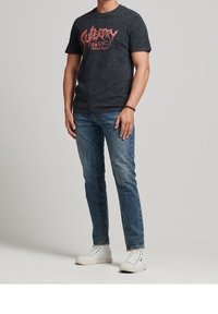 Camiseta gráfica negra con el texto "Superdry Japan Tour", combinada con jeans de mezclilla azul y zapatillas altas blancas. Diseño simple, ajuste casual.