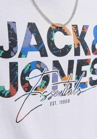 Weißes T-Shirt mit kräftigem schwarzen Text "JACK & JONES" und "Essentials" in buntem Spritzdesign, ergänzt durch eine silberne Kettenhalskette.