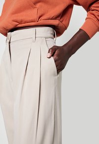 Ljust beige veckade byxor med hög midja och sidfickor. Tillverkade av texturerad tyg har midjebandet en knappstängning.