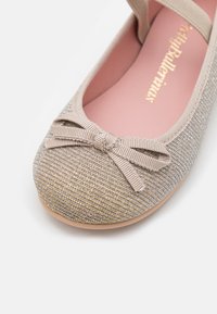 Ballerina beige con un esterno in tessuto texturizzato, fili metallici e un piccolo fiocco sulla punta. Fodera interna rosa e suola flessibile.