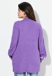 Lila gestrickter Pullover mit lockerer Passform, langen Ärmeln und gerippten Details, getragen über hellblauen Jeans. Weiche Textur sichtbar.