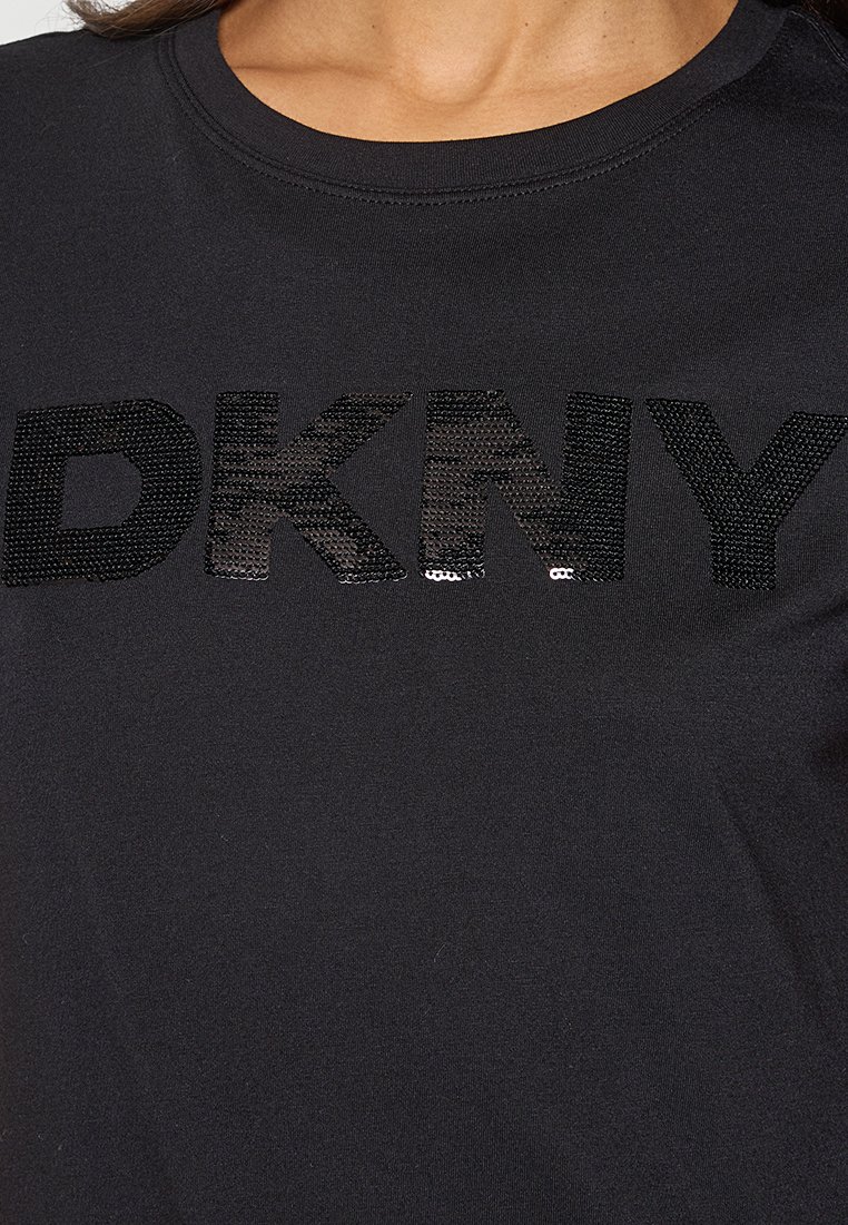 Camiseta negra con un audaz logo de lentejuelas negras texturizadas "DKNY" en el pecho. Tejido suave con cuello redondo.