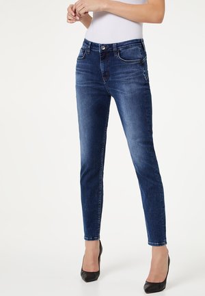 Femme portant un jean skinny bleu foncé et des talons noirs à bout pointu, debout devant un fond blanc uni.