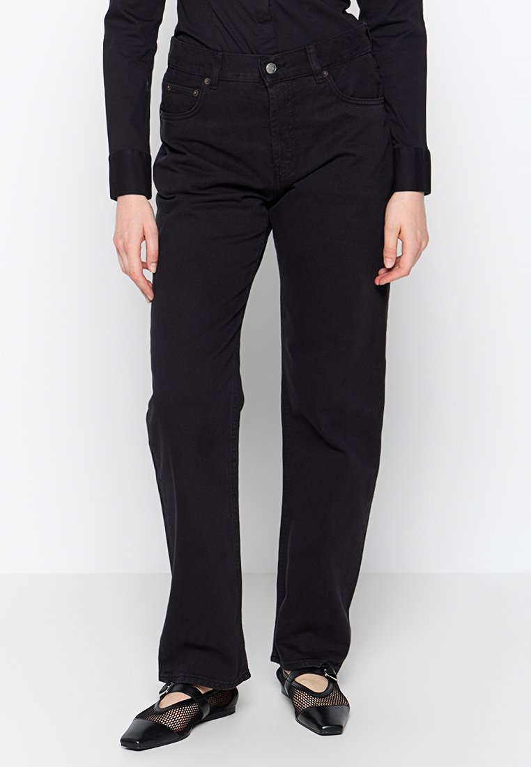 Zwarte denim broek met een rechte pijp, vijf zakken en metalen knoopsluiting. Gedragen met zwarte schoenen met gaasaccenten.