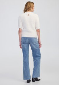 Witte, donzige, mouwloze trui met een sleutelgat detail aan de achterkant, gecombineerd met medium-wash flared jeans en zwarte haklaarzen.