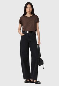 Camicia marrone a maniche corte, jeans neri a gamba larga a vita alta, cintura nera con accessori dorati e scarpe nere basse, mentre tiene una borsa a tracolla nera.