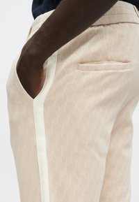 Beige mønstrede bukser med et geometrisk design, med hvit sidestripe, skrå lomme og detalj på baklommen.