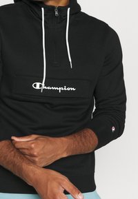 Felpa nera con zip bianca e coulisse. Caratterizzata da una tasca frontale con logo "Champion" stampato e polsini a costine con accento del logo.