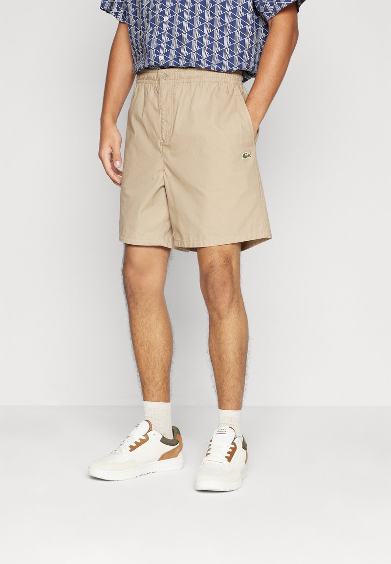 Beige shorts laget av bomull med elastisk midje og sidelommer, utstyrt med en liten grønn Lacoste-logo. Kombinert med hvite joggesko.