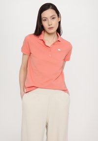 Koralo spalvos polo marškiniai su apykakle, dviejų mygtukų užsegimu ir mažu logotipu. Derinami su šviesiais beige spalvos kelnėmis, turinčiomis laisvą siluetą.