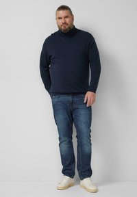 Marineblauer Pullover mit hohem Kragen, kombiniert mit blauen Jeans und weißen Turnschuhen. Der Pullover hat eine weiche Textur und eine taillierte Silhouette.