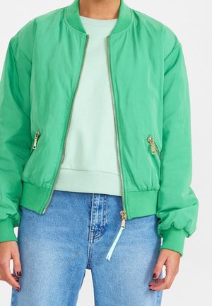 Personne portant une veste bomber verte par-dessus un pull vert clair et un jean bleu, debout avec les mains détendues le long du corps.