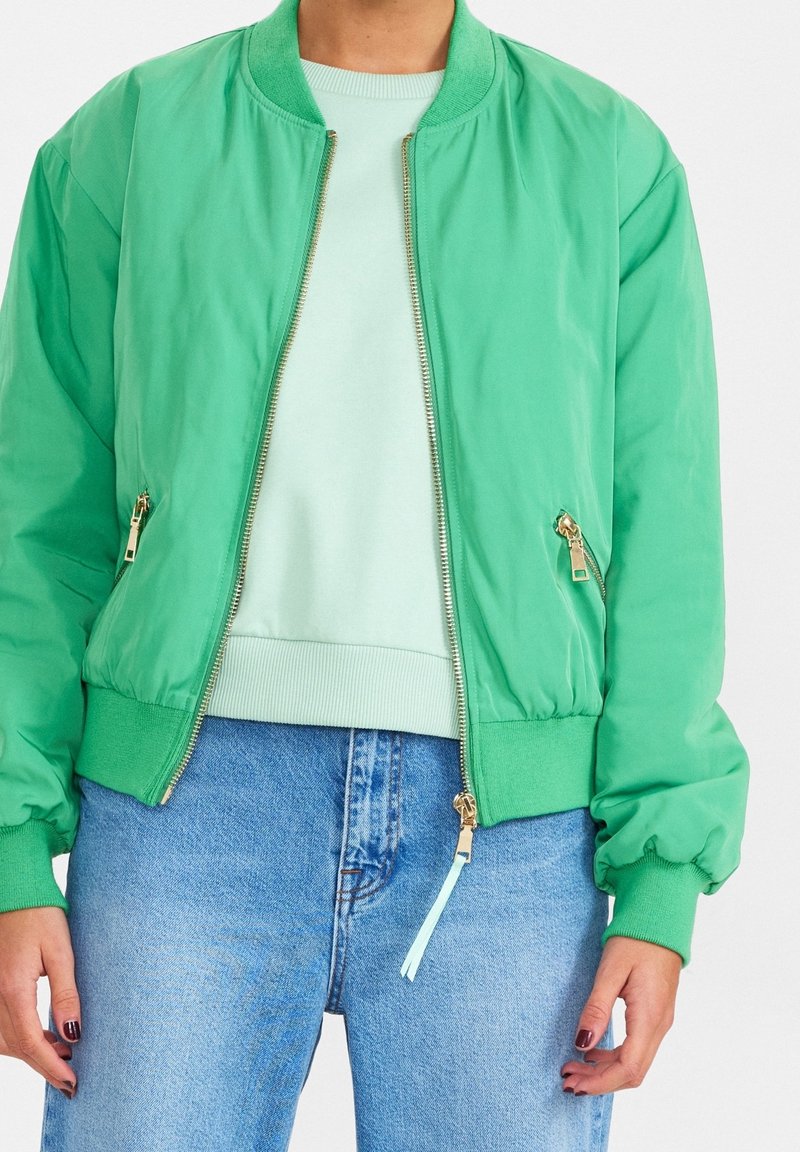 Personne portant une veste bomber verte par-dessus un pull vert clair et un jean bleu, debout avec les mains détendues le long du corps.
