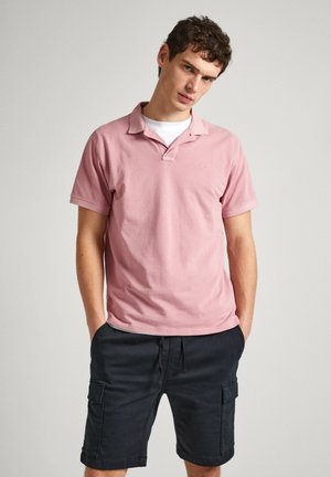 Polo shirt rosa chiaro con colletto a bottoni, abbinato a pantaloni cargo blu navy con tasche e cintura con cordoncino. Modello in piedi con le mani in tasca.