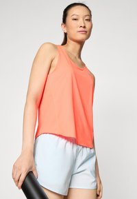 Débardeur en corail vif, fabriqué en tissu respirant avec une coupe ample. Associé à un short bleu clair, tenant un tapis de yoga noir.