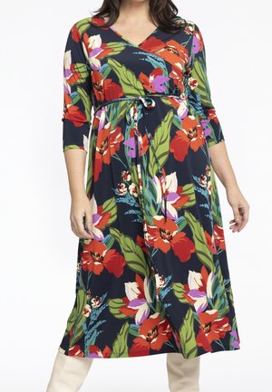 Kobieta ubrana w granatową sukienkę kopertową z czerwono-biało-fioletowym kwiatowym wzorem i zielonymi liśćmi, z rękawami do łokcia i wiązaniem w pasie.