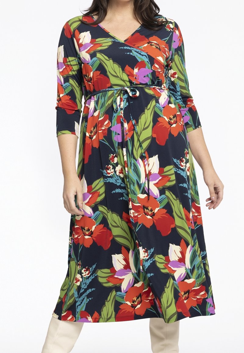 Femme portant une robe portefeuille bleu marine avec un motif floral rouge, blanc et violet accompagné de feuilles vertes, dotée de manches trois-quarts et d'une ceinture à la taille.