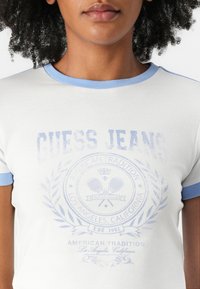 Valge lühikeste varrukatega T-särk helesinise soonikuga kaeluses, millel on tuhm "Guess Jeans" graafika ja dekoratiivne ümmargune logo.