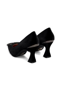 Zapatos de ante negros de tacón medio con tacón acampanado y detalle decorativo de rhinestones en la puntera, mostrados desde los ángulos trasero y lateral.