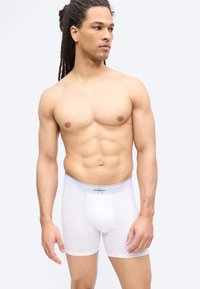 Μυώδης άνδρας με περουβιανά dreadlocks, φορώντας λευκά εσώρουχα Calvin Klein boxer, στέκεται μπροστά σε απλό ανοιχτό φόντο, κοιτάζοντας προς τα αριστερά του.