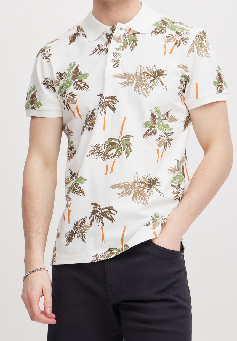 Witte polo met korte mouwen, versierd met een palmbomenpatroon in groen, bruin en oranje, met een klassieke kraag en een rechte zoom.