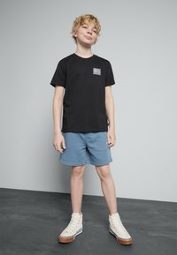 Svart t-skjorte med korte ermer som har et fargerikt mønster på venstre bryst, i følge med lyseblå shorts og hvite høyhælte sneakers.
