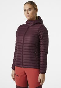 Bordový péřový kabát s kapucí, s vodorovným prošívaným designem, plným předním zipem a elastickými manžetami. V kombinaci s oranžovými kalhotami.