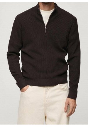 Mann trägt einen dunkelbraunen, gerippten Viertel-Zip-Pullover über einem weißen Hemd, kombiniert mit hellbeigen Hosen, steht mit einer Hand in der Tasche.