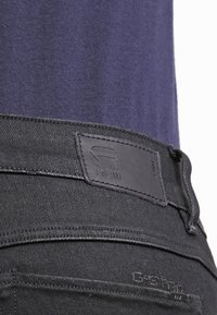 Jeans de mezclilla negra con una etiqueta texturizada en la cintura. Presenta un diseño liso y detalles de costura sutiles, con una parte superior azul marino parcialmente visible.