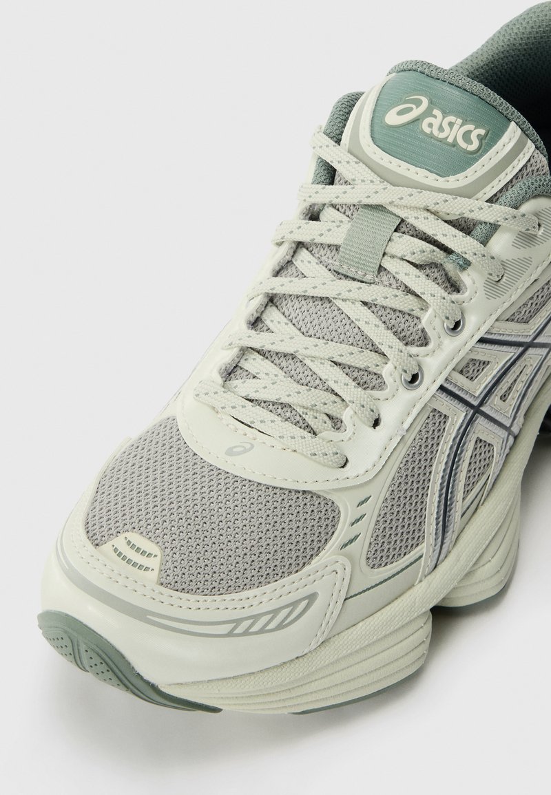Chaussure de sport gris clair avec tige en maille, accents verts et semelle texturée. Présente des lacets, un logo et un design en couches.