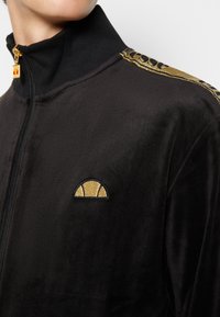 Svart zip-up jacka i sammet med hög krage. Har en guld broderad logotyp på bröstet och ett guldaccent längs axeln.