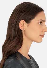 Zwarte studoorbel weergegeven op het oor van een model, met een ronde vorm en een gladde textuur. Het model heeft lang bruin haar en draagt een zwarte top.