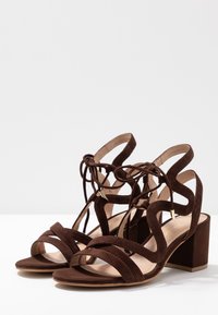 Sandalias de tacón alto en ante marrón con un diseño de tiras, punta abierta y tacón cuadrado, con detalles de cordones y acentos metálicos.