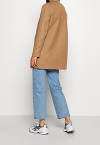 Lang, kamelfarvet frakke lavet af tekstureret stof, åben foran, kombineret med lyseblå jeans med lige ben og sporty hvide sneakers.