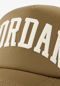 Brūna cepure ar izteiktu baltu "JORDAN" uzrakstu. Izgatavota no teksturētas auduma ar izliekto samtu un izšūtiem akcentiem apakšpusē.