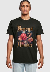 Sort bomulds t-shirt med et blomstermønster i klare farver og guldskrift, der siger "Beyond Humble." Enkel crew neck design.